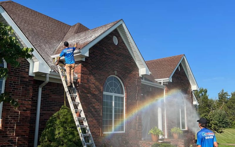 professional standing on ladder washing roof of brick home, other professional soft washing siding